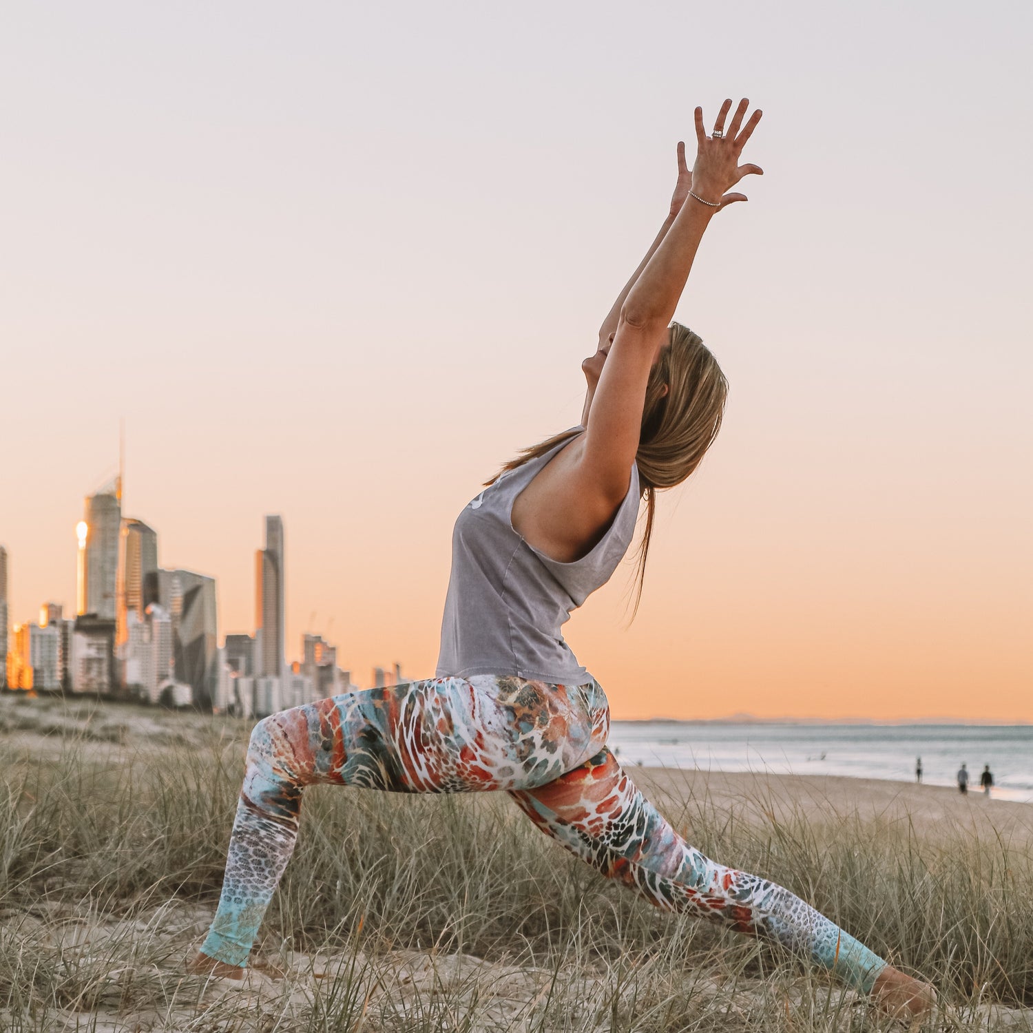 Person practicing yoga on a grassy field with a city skyline and beach in the background during sunset.